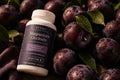 Rytma Magnesium Glycinate bottle resting among dark purple plums and wet green leaves in low evening light.